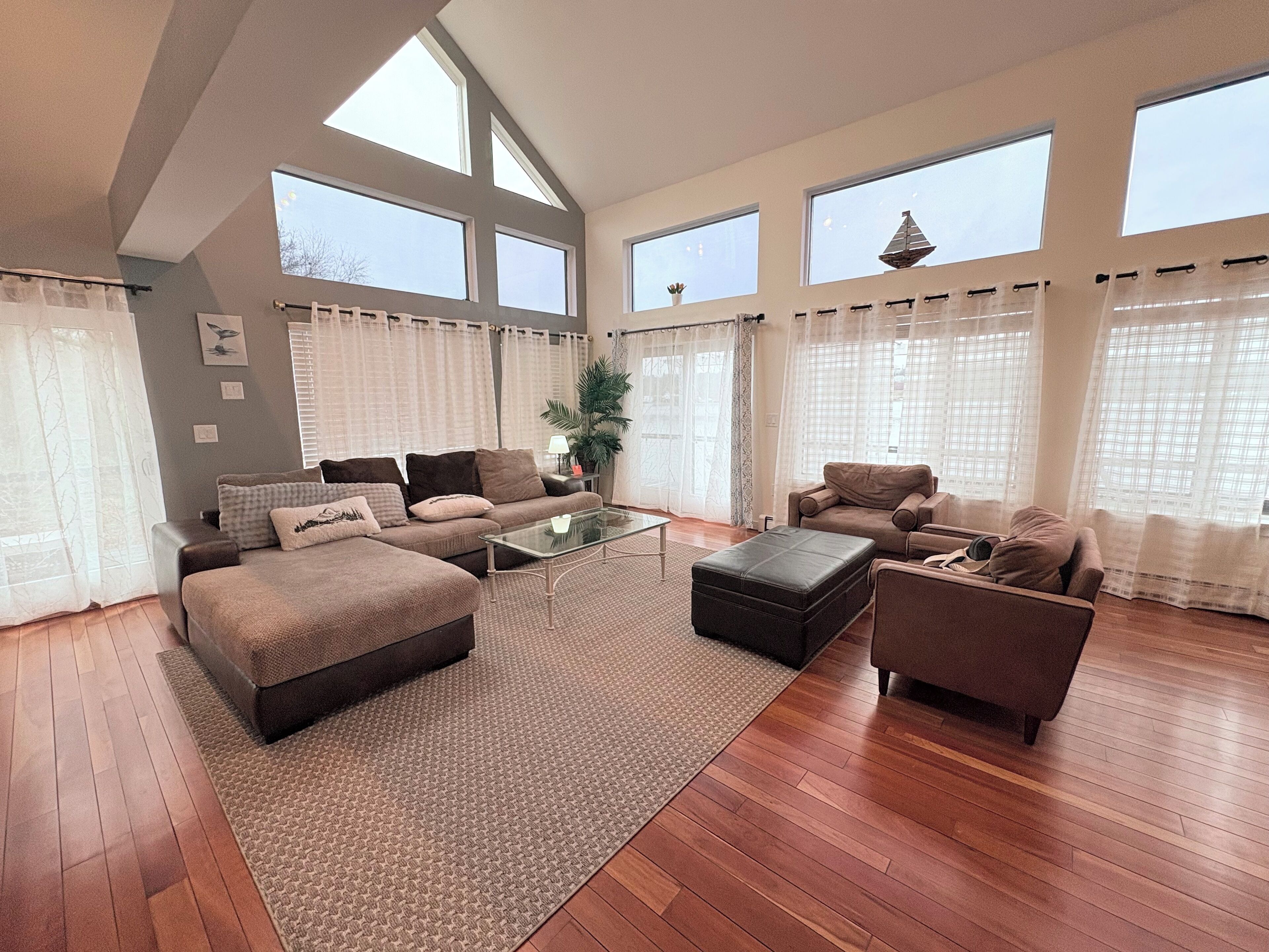 A spacious living room with a large sofa set, a coffee table, and a small side table. There are large windows with sheer curtains, a potted plant, and a decorative item on the windowsill.