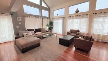 A spacious living room with a large sofa set, a coffee table, and a small side table. There are large windows with sheer curtains, a potted plant, and a decorative item on the windowsill.