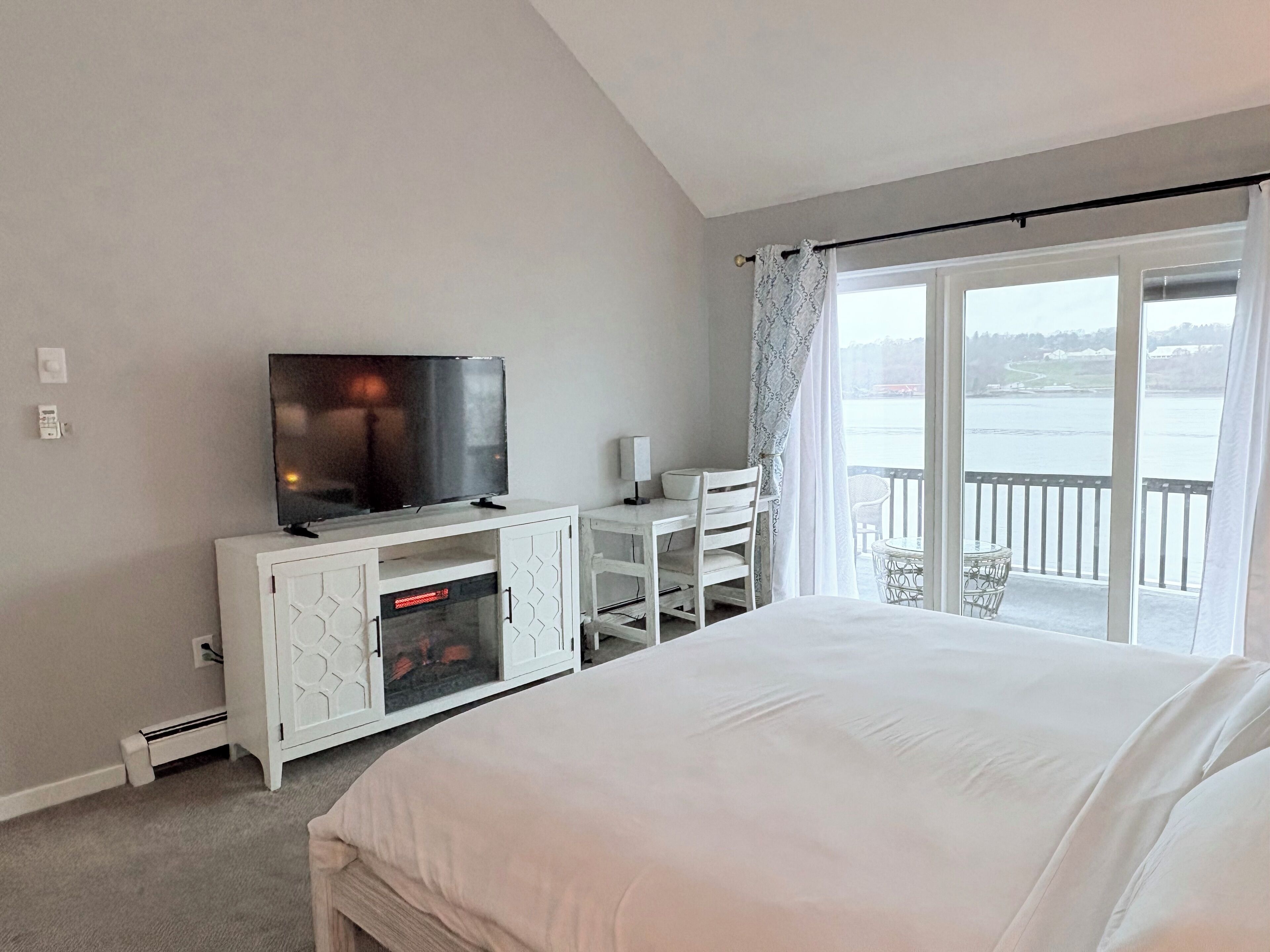 A hotel room with a bed, a TV on a white cabinet, a desk with a chair, and a balcony with a view of the water.