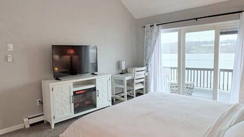 A hotel room with a bed, a TV on a white cabinet, a desk with a chair, and a balcony with a view of the water.