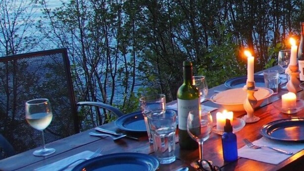 Une table dressée pour un repas, avec des bouteilles de vin, des verres et des bougies, donnant sur une étendue d’eau.
