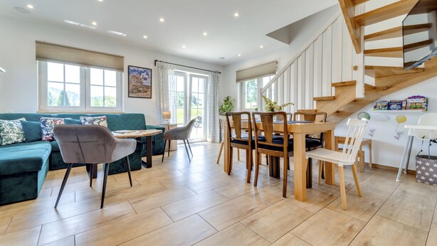 A spacious living area with a wooden staircase, a dining table with chairs, a sofa, and a large window providing natural light. - Siglanów by Interhome (Wisla)