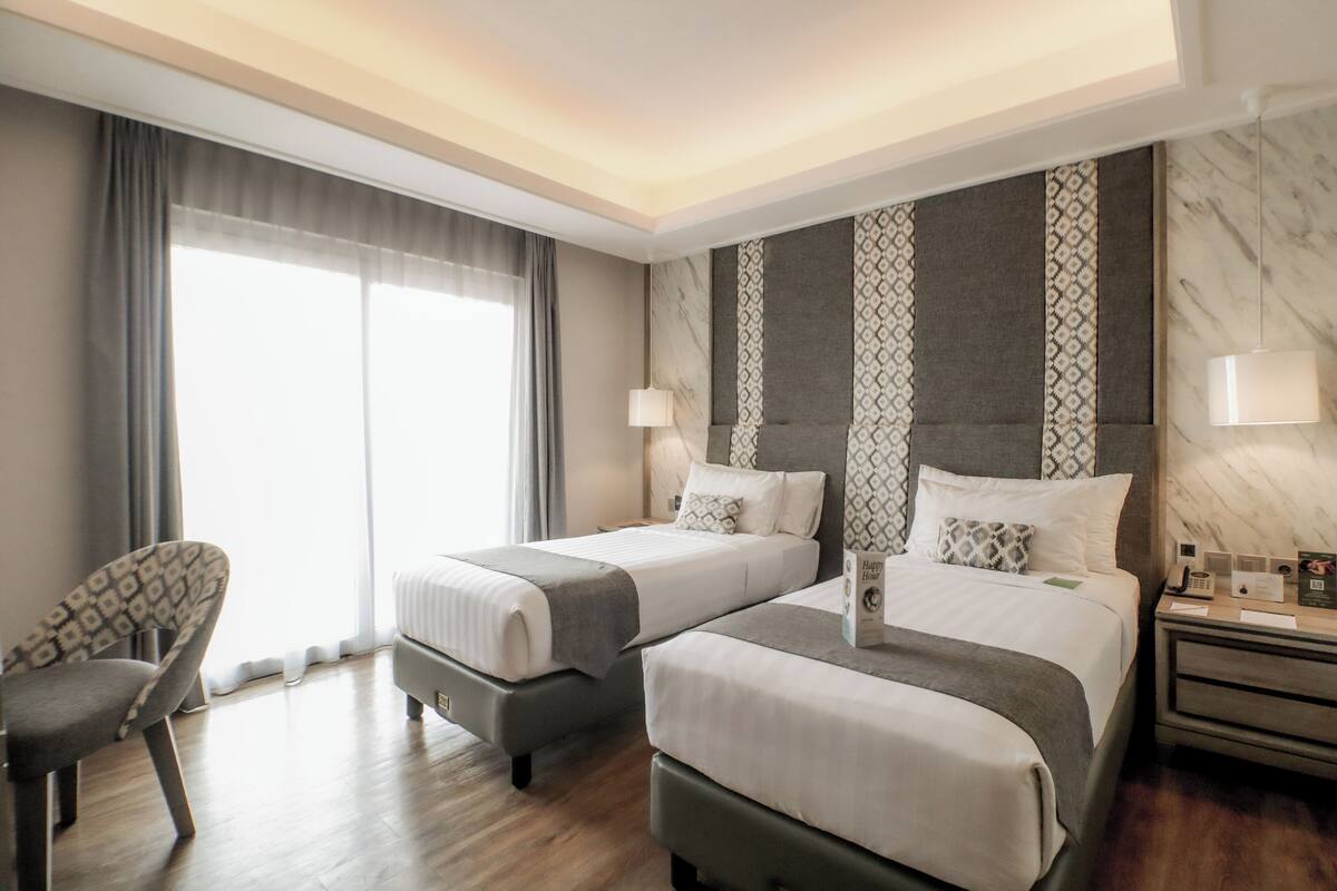 A hotel room with two beds, a chair, a nightstand with a lamp, and a large window with curtains.
