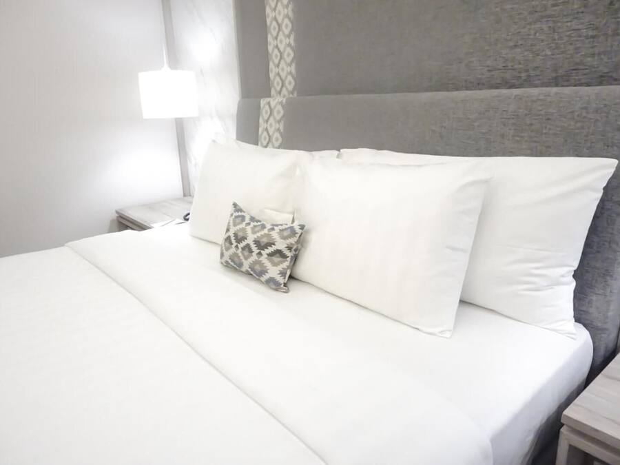 A neatly made bed with white linens and a patterned pillow, a bedside table with a remote control, and a wall with a textured pattern.