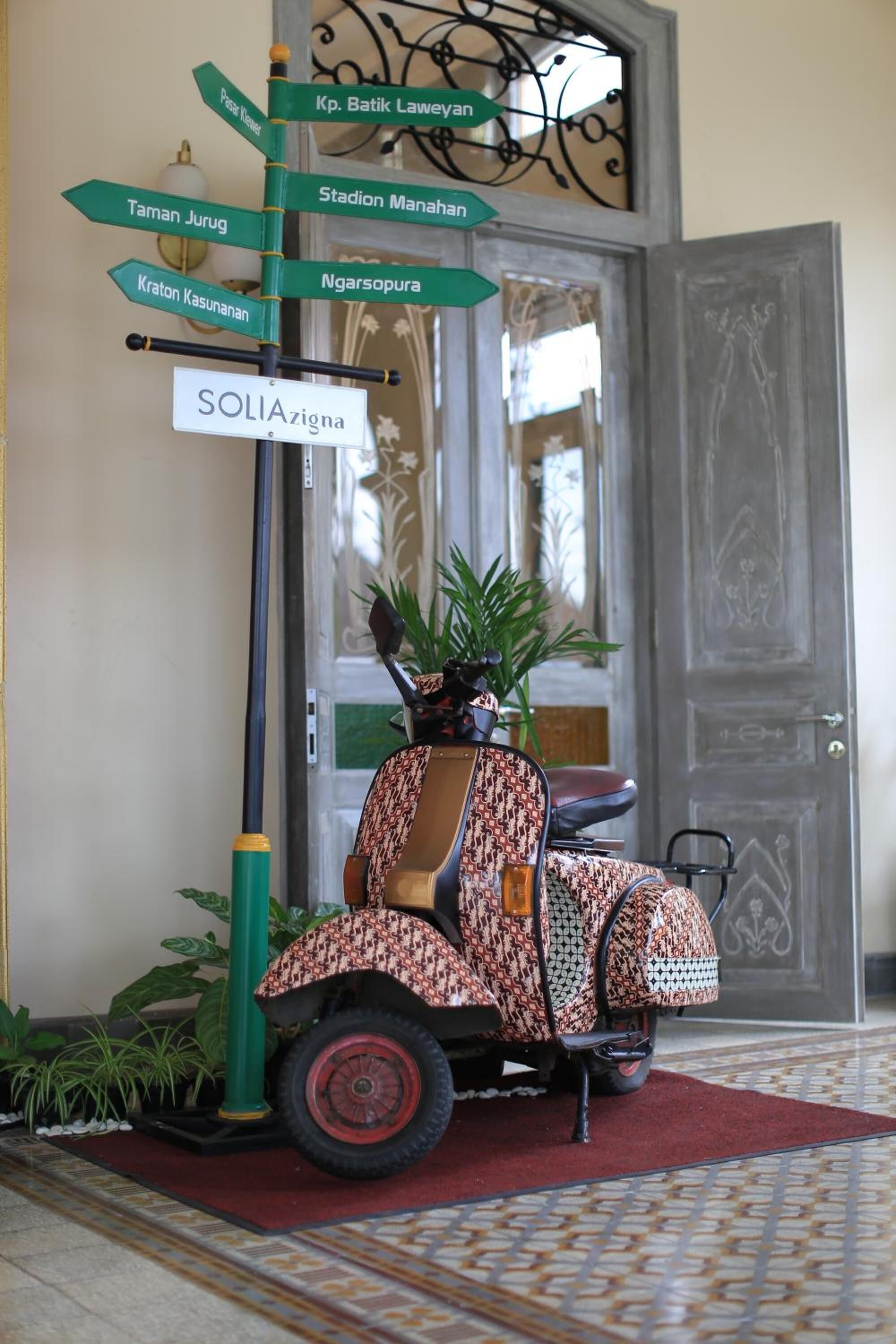 Een klassieke scooter staat geparkeerd voor een deur met een bord waarop verschillende bestemmingen zijn aangegeven.
