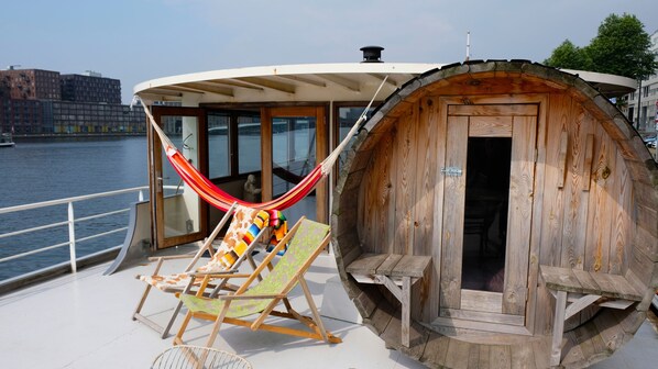 Una cabaña de madera con un ventanal grande y una puerta, una hamaca y una silla de descanso en la cubierta de un barco.