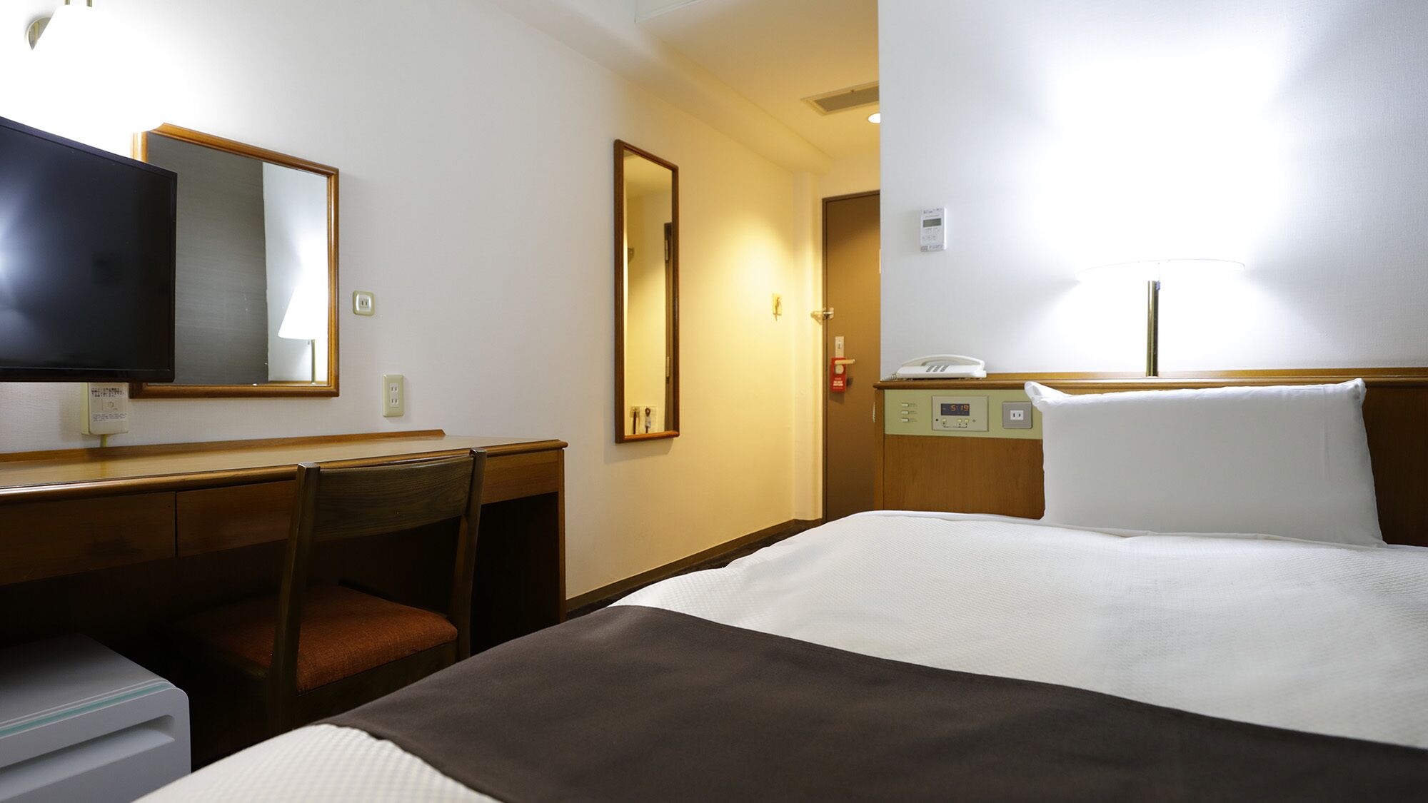 A hotel room with a bed, a desk, a chair, a TV, a mirror, a bedside table with a phone, and a wall clock.