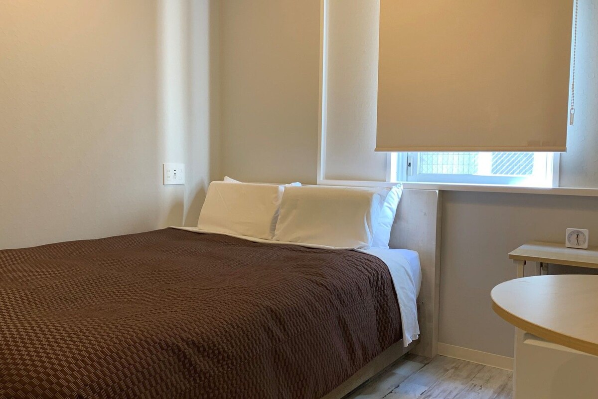 A small, neatly arranged bedroom with a single bed, a bedside table, and a window with blinds.