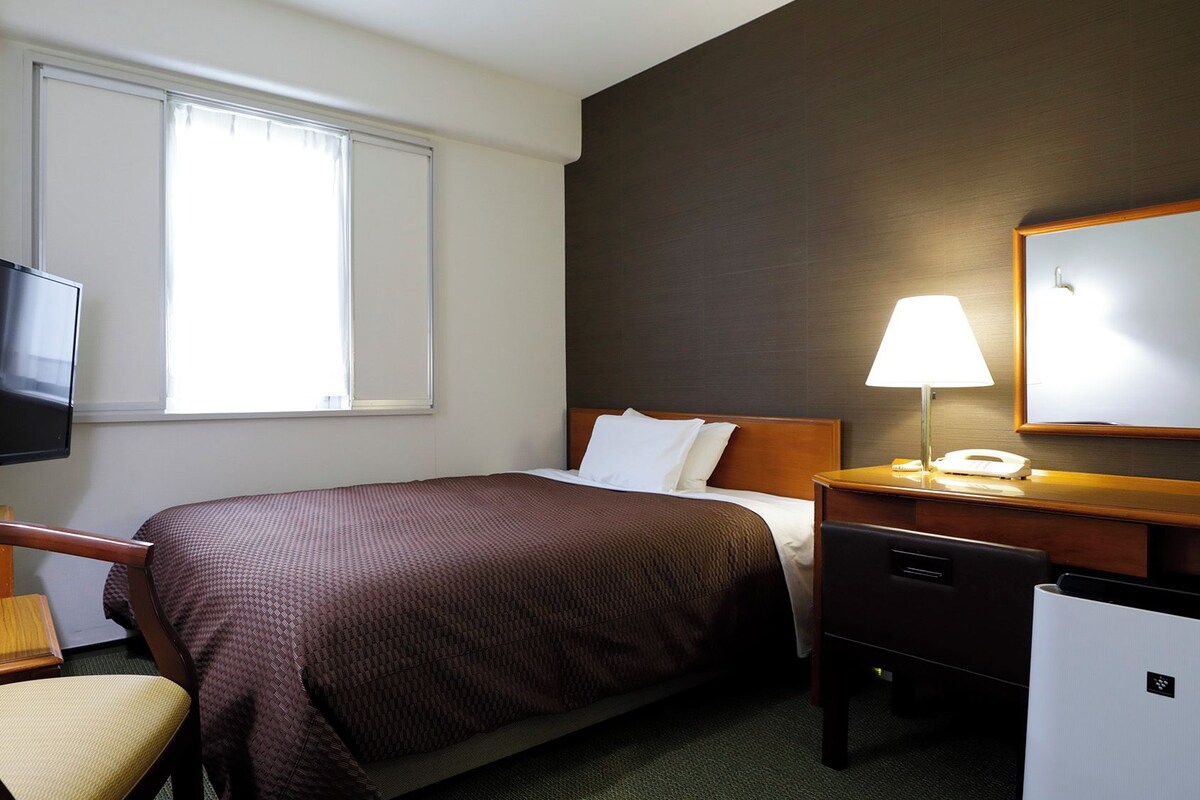 Una habitación de hotel con cama, escritorio con lámpara, silla, televisor y ventana.