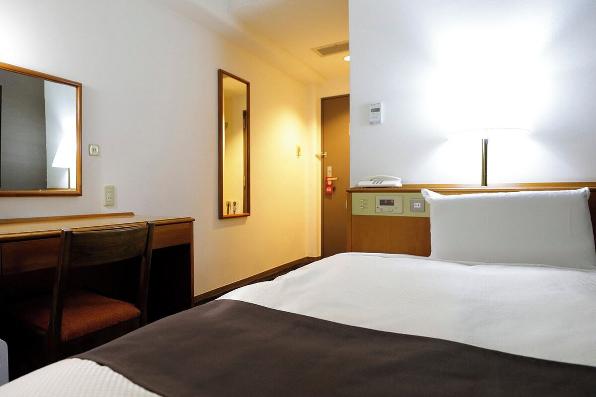 Habitación de hotel con cama, escritorio con silla, televisión, espejo, mesita de noche y termostato.