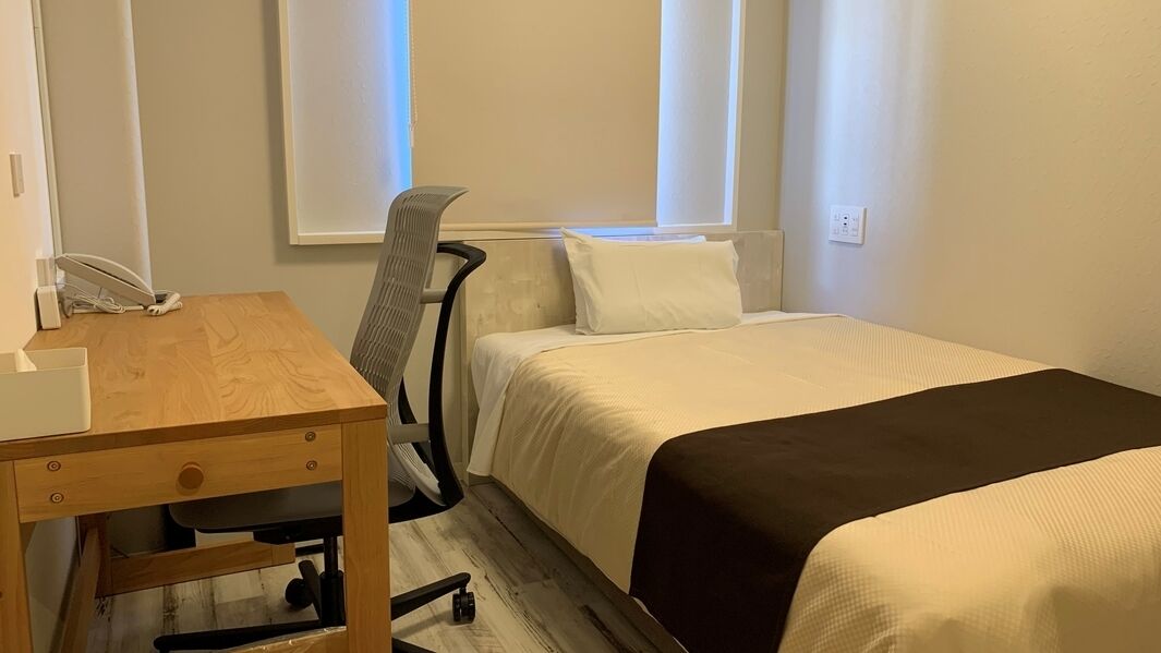 A hotel room with a wooden desk, a chair, a bed with white and brown bedding, and a window with blinds.