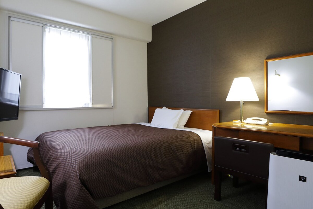 Habitación de hotel con cama, escritorio con lámpara, silla, televisor y refrigerador.
