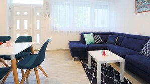 A modern living room with a blue sofa, a white coffee table, and a dining table with chairs. There are two pendant lights above the sofa and a large window with sheer curtains. - Vacation home Pixie by Interhome (Balatonoszod)
