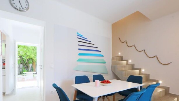 A modern dining area with a white table and blue chairs, a staircase with wooden steps, and a wall with abstract art. - Vacation home Pixie by Interhome (Balatonoszod)