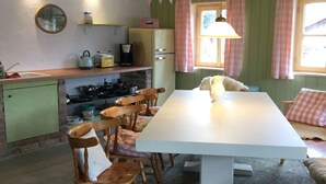 A dining area with a long table, wooden chairs, a rug, and a kitchen in the background. - Ferienwohnung Immergrün (Görisried)