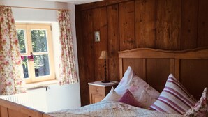 A wooden bed with a quilt, a nightstand with a lamp, and a window with floral curtains. - Ferienwohnung Immergrün (Görisried)