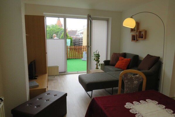 A room with a sofa, ottoman, and a table with a doily. A television is on a stand, and a plant is on the floor. - Apartment am Klinikum - Apartments am Klinikum (Dessau-Roßlau)