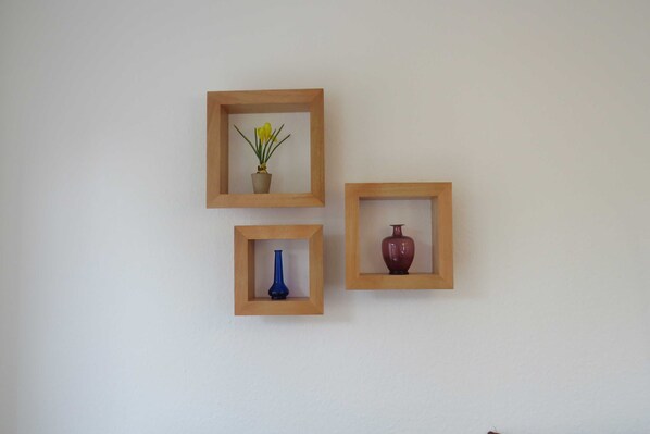 Three square wooden frames on a wall, each containing a different object: a potted plant, a blue vase, and a red vase.