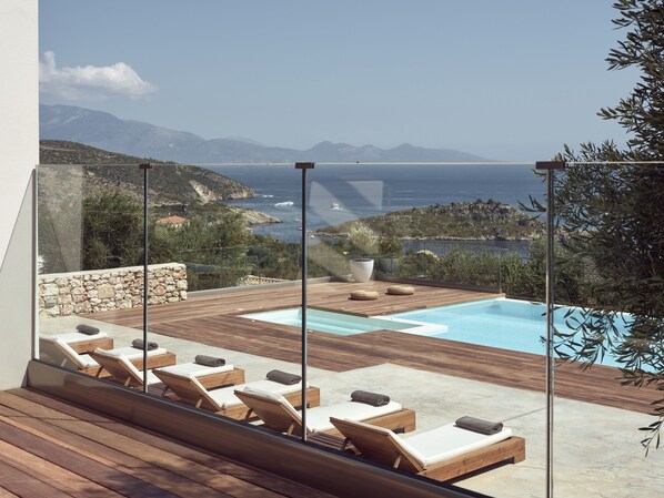 A modern outdoor pool area with wooden loungers and a view of the sea and mountains.
