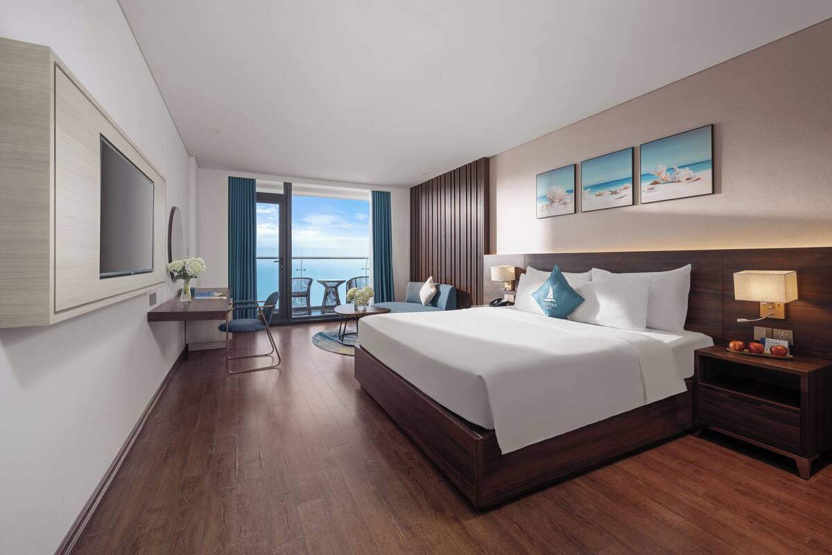 A modern hotel room with a large bed, a desk with a chair, a view of the ocean, and a balcony.