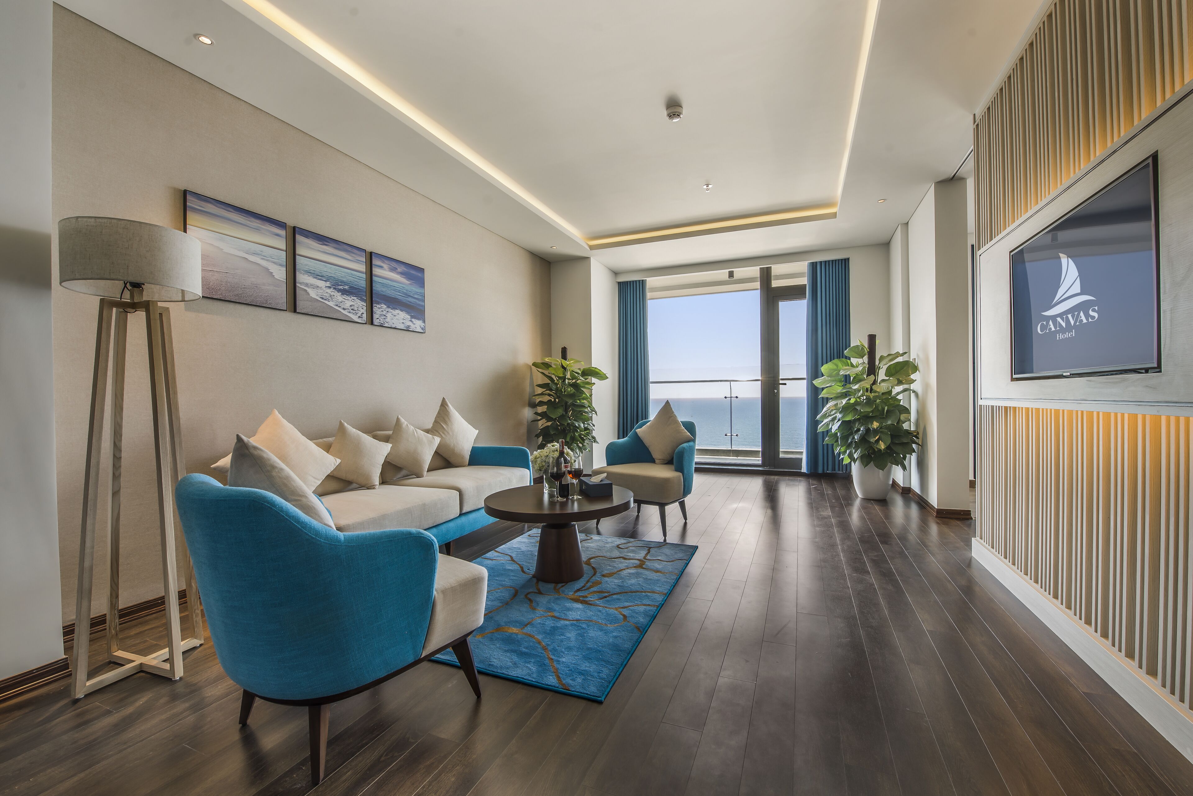 A modern hotel lobby with a sofa, chairs, a coffee table, a floor lamp, and a TV screen.