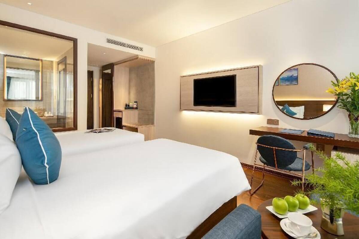 A hotel room with a bed, a desk with a chair, a TV, a mirror, and a plant.