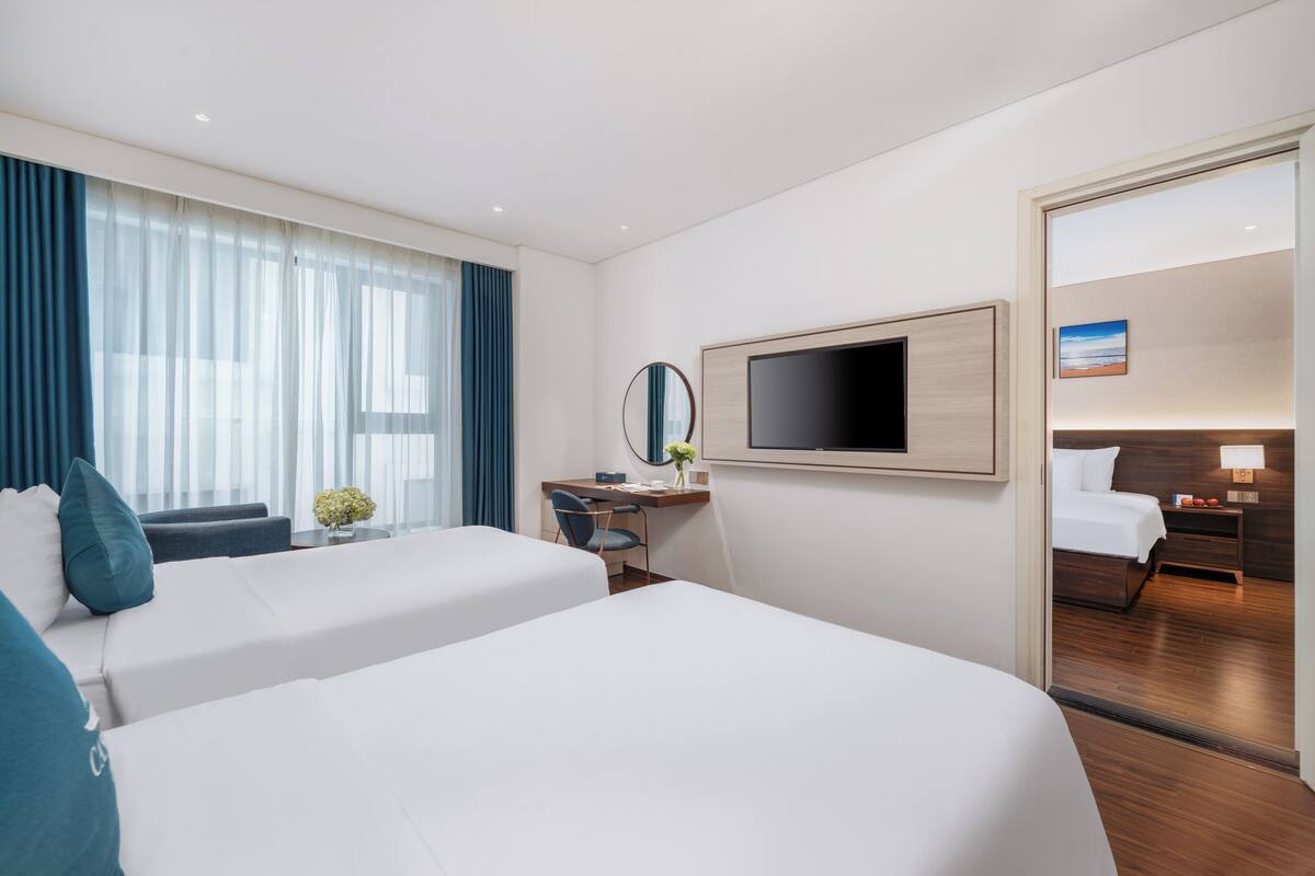A hotel room with two beds, a desk, a TV, and a view of the room through a glass door.