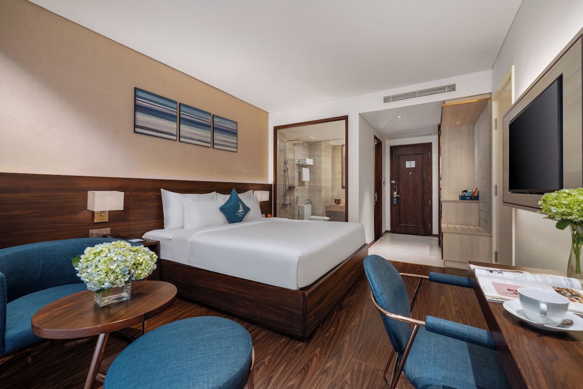 A hotel room with a bed, a seating area with a table and chairs, a TV, and a bathroom visible through an open door.