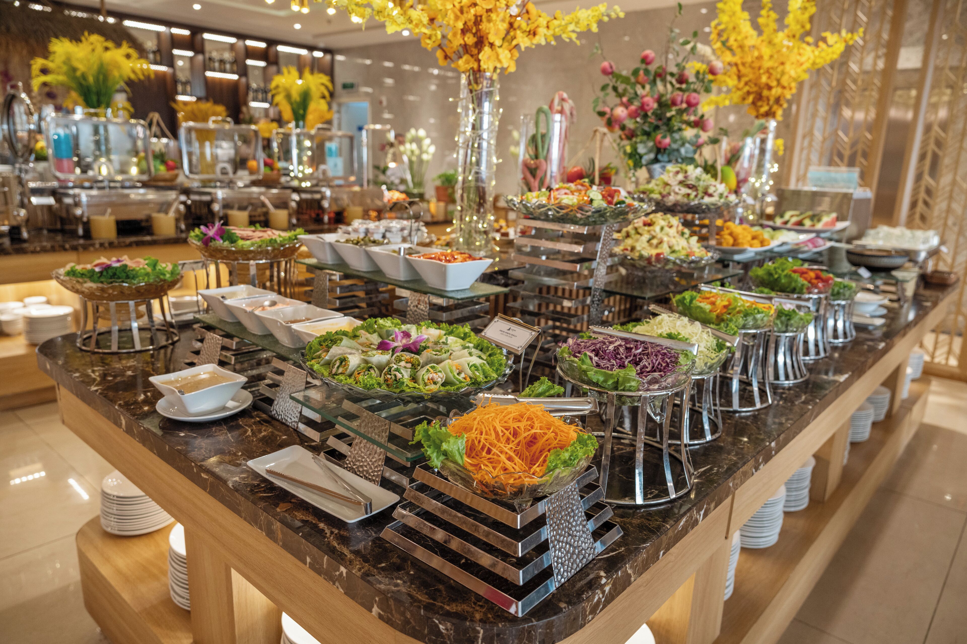 A buffet with various food stations, including salads, vegetables, and other dishes, arranged on a marble counter with a wooden base.