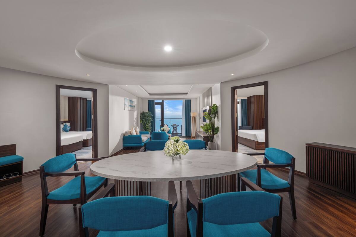 A modern hotel room with a round dining table, blue upholstered chairs, a marble-top table, and a view of the sea through a large window.