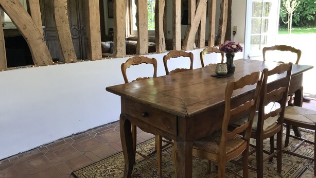 Een houten eettafel met zes stoelen, een vaas met bloemen en een vloerkleed op een betegelde vloer.