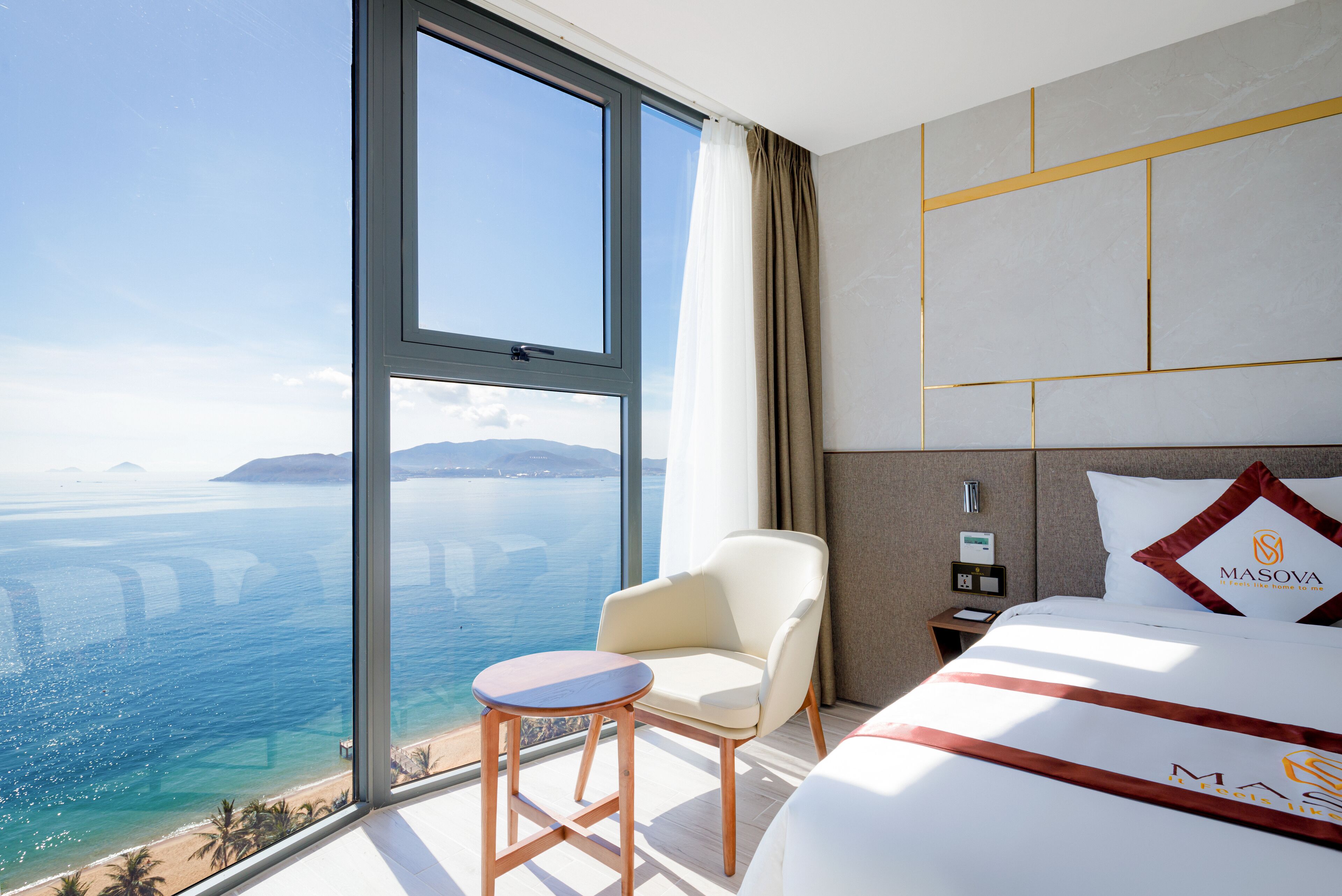 Una camera d'albergo con una grande finestra, un letto con cuscino a marchio, un tavolino e un comodino con lampada.