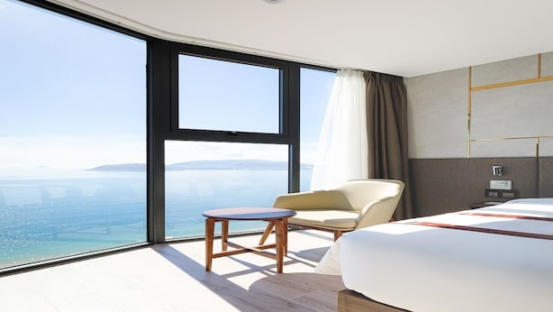 A modern hotel room with a large window offering a sea view, a bed, a chair, and a small table. - Masova Hotel (Nha Trang)