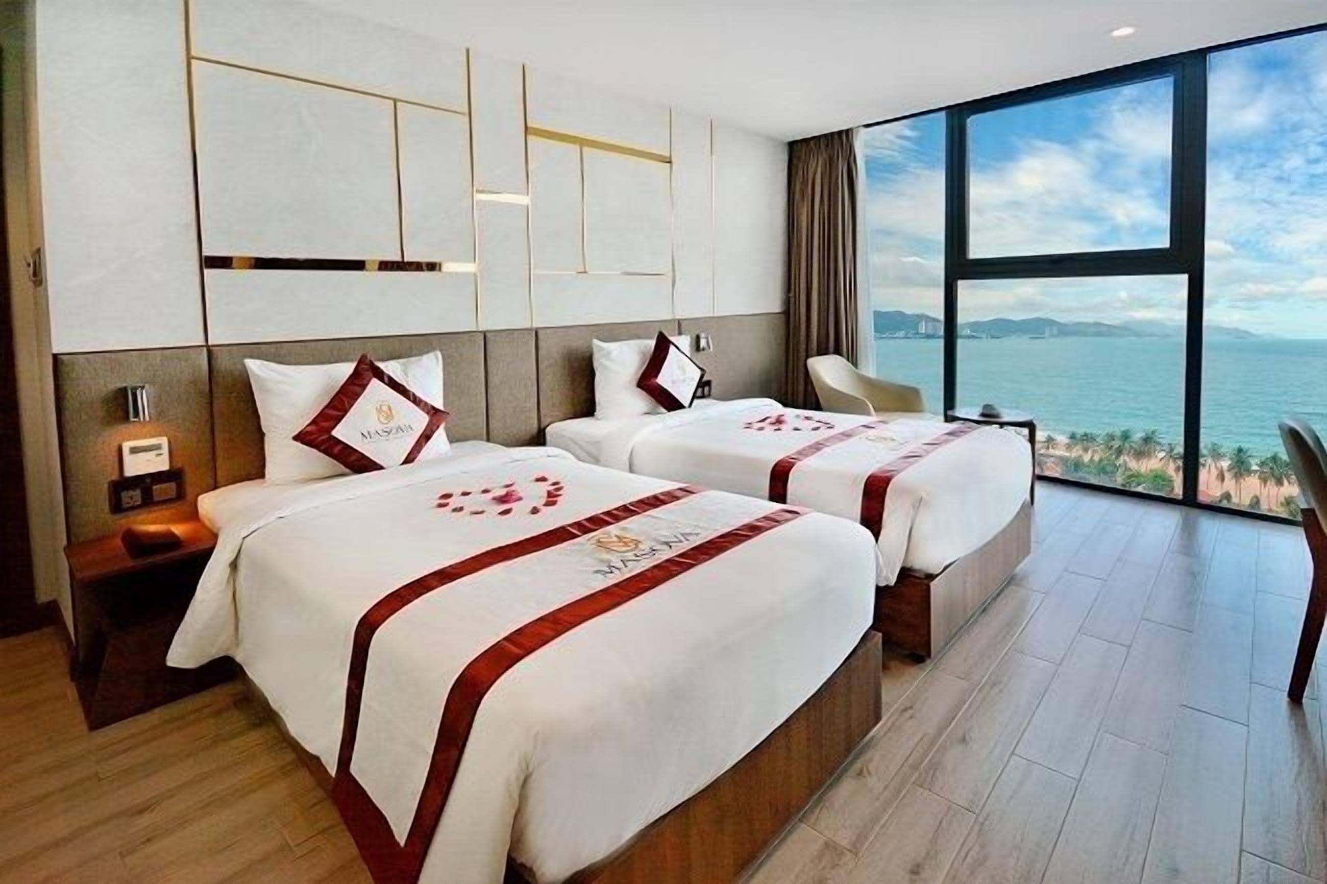 A hotel room with two beds, a nightstand, a lamp, and a large window with a view of the sea.