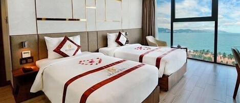 A hotel room with two beds, a nightstand, a lamp, and a large window with a view of the sea.