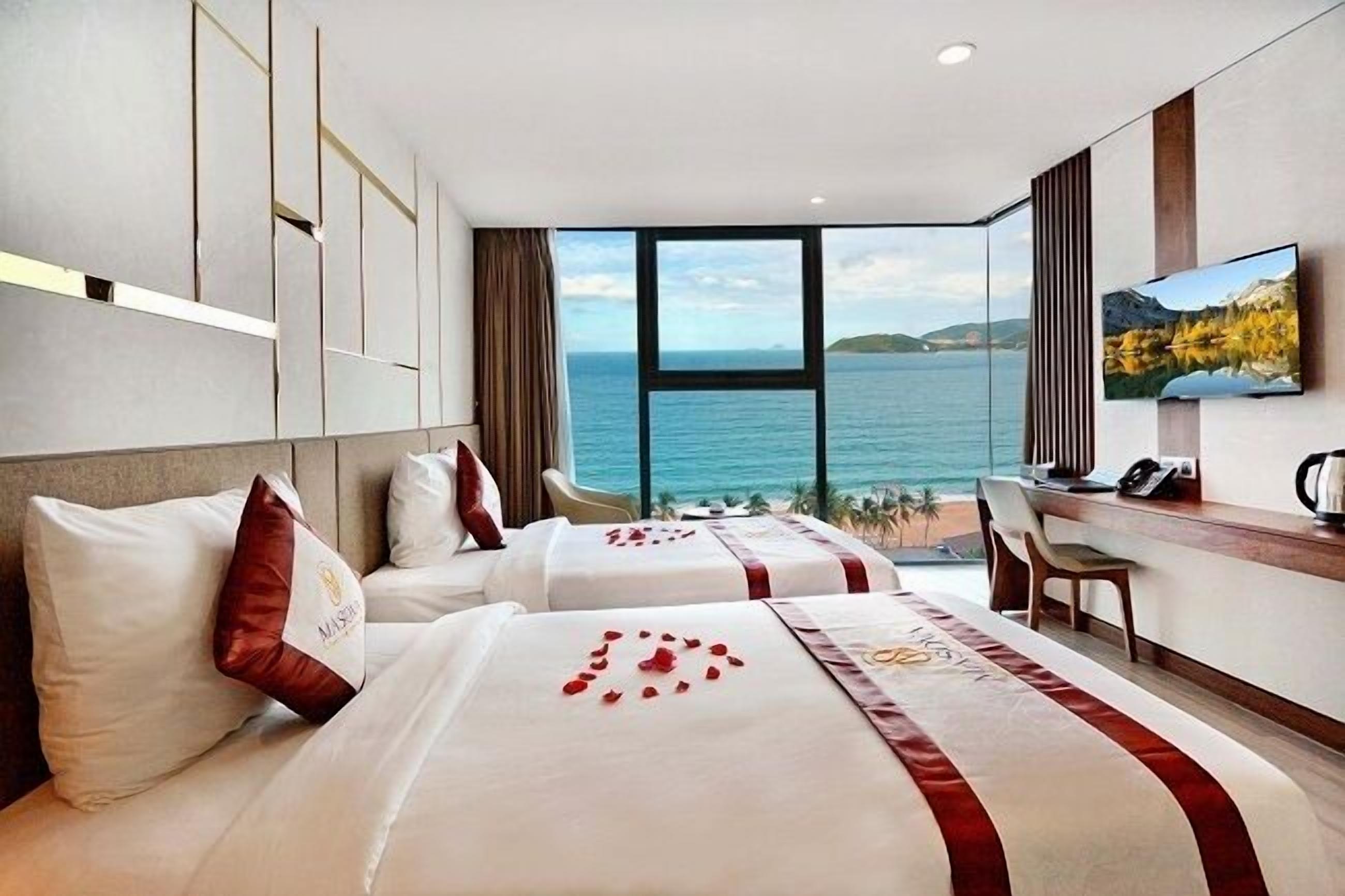 A hotel room with two beds, a desk, a TV, and a view of the sea.
