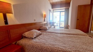 A hotel room with two beds, a wooden headboard, a nightstand, a lamp, and a view of the beach through a sliding door.