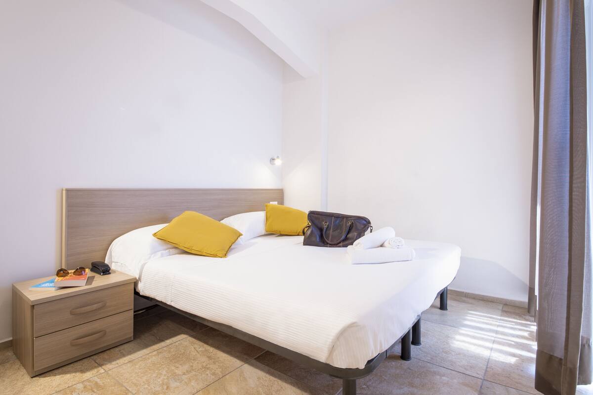 A neatly made bed with white bedding and two yellow pillows, a bedside table with a book and a phone, and a brown handbag on the bed.