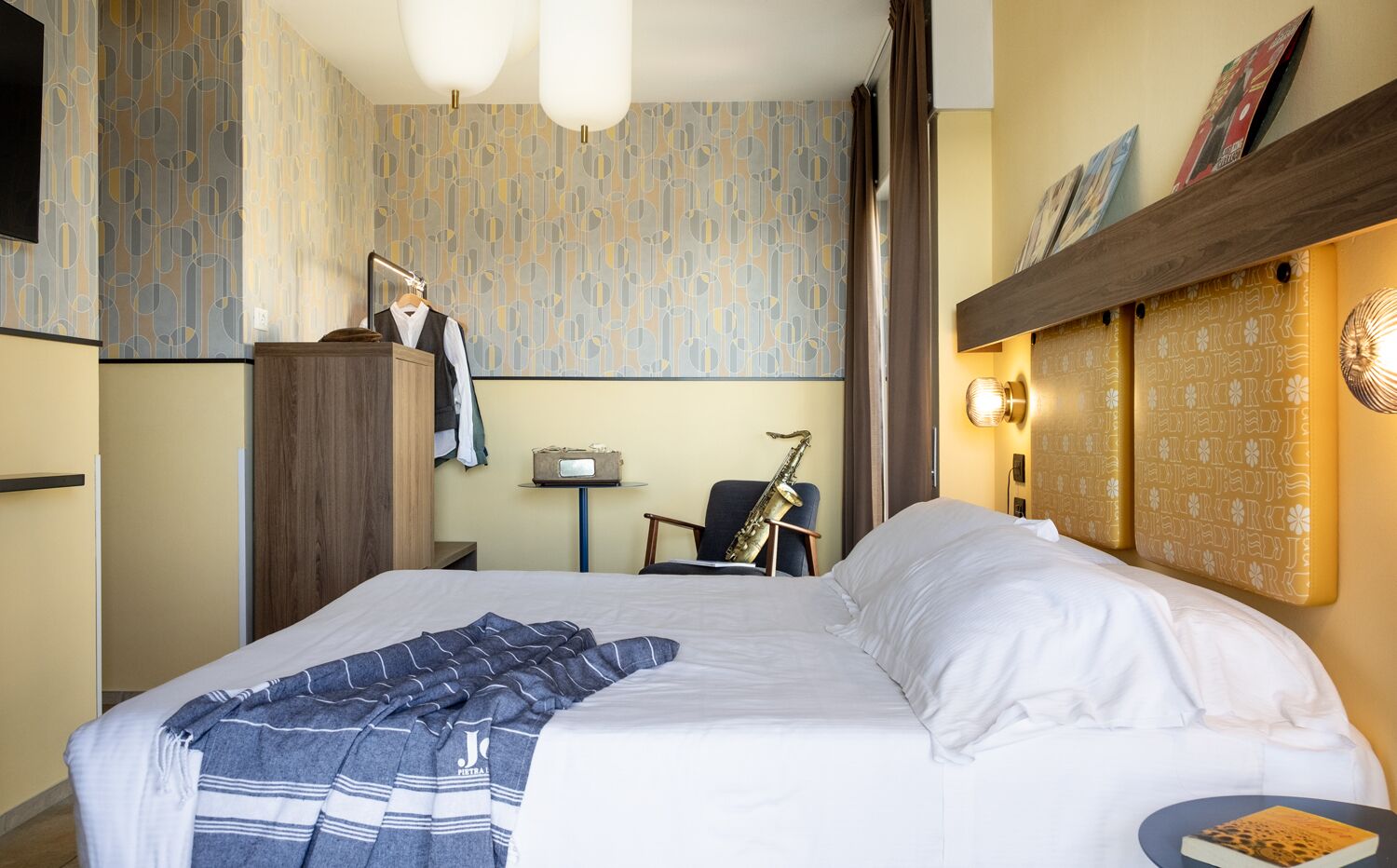 A hotel room with a large bed, a desk with a chair, a saxophone, and a small table with a book.