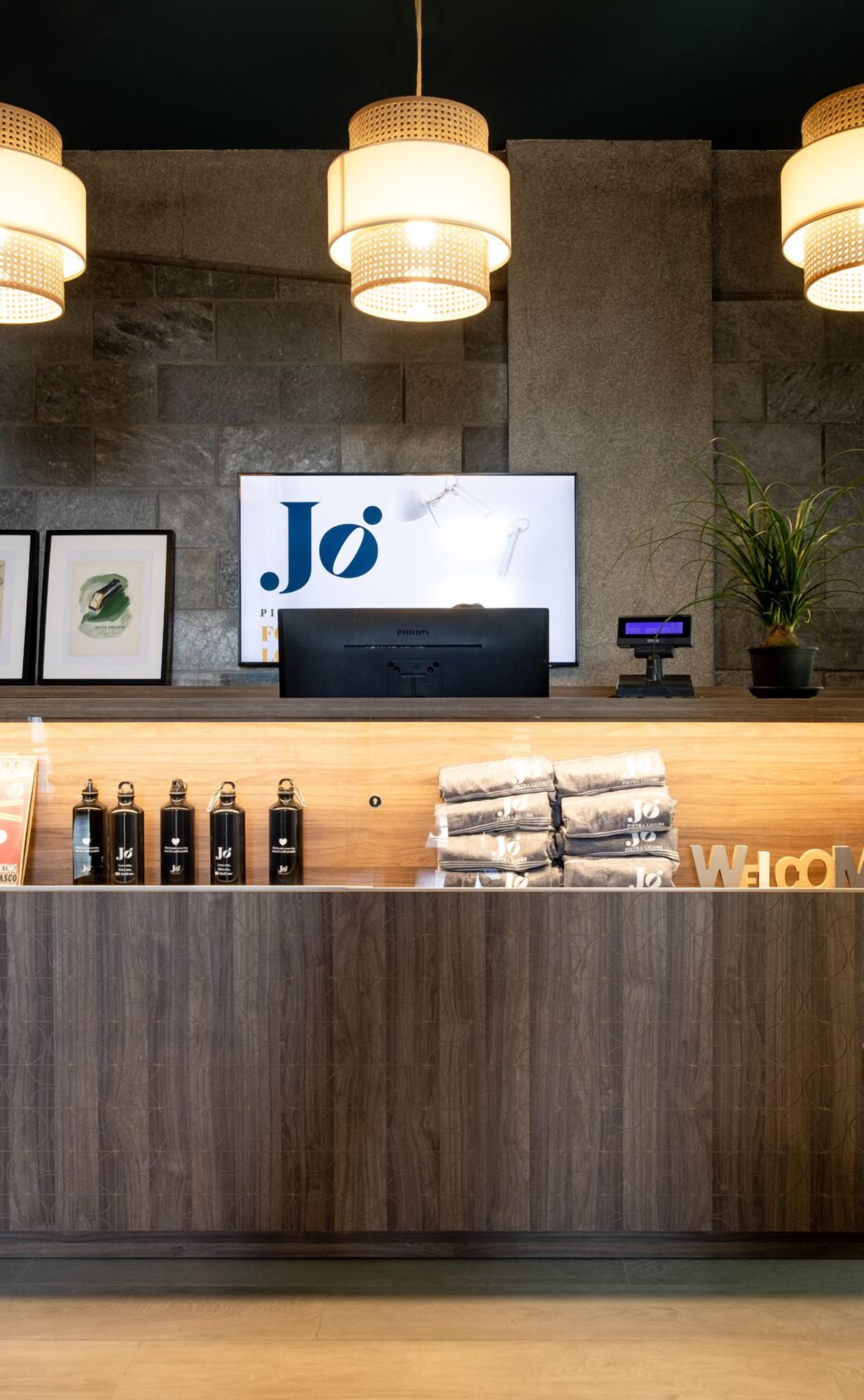 Un'area di accoglienza con un bancone in legno, uno schermo televisivo che mostra il logo "Jo" e tre lampade pendenti a sospensione.