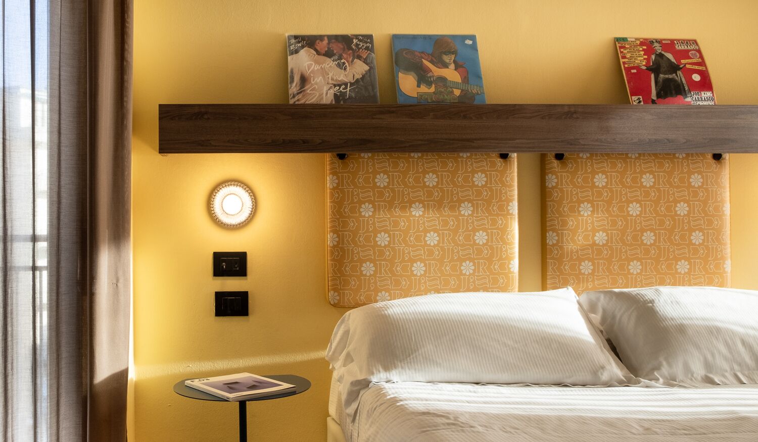 A hotel room with a bed, a small table, a wall-mounted light, and decorative items on a shelf above the bed.
