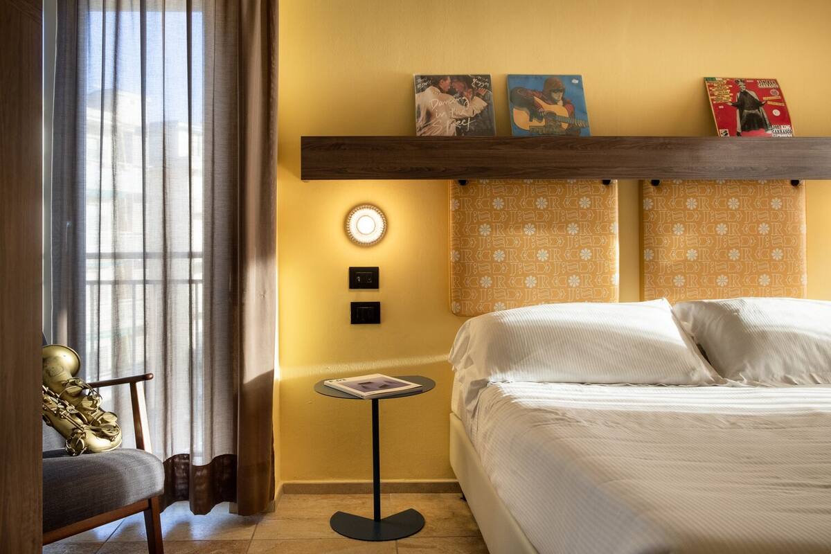 A hotel room with a bed, a small table, a chair, a window with curtains, and wall decorations.