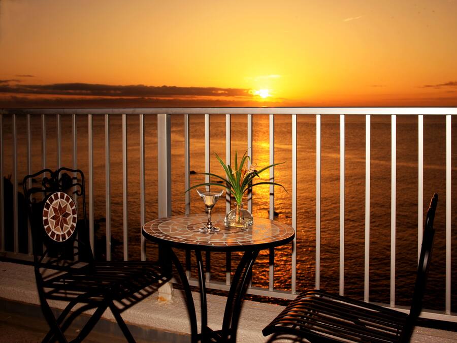 Una mesa redonda con sillas y un florero de vidrio con una planta, frente a una vista del atardecer en un balcón con barandilla.