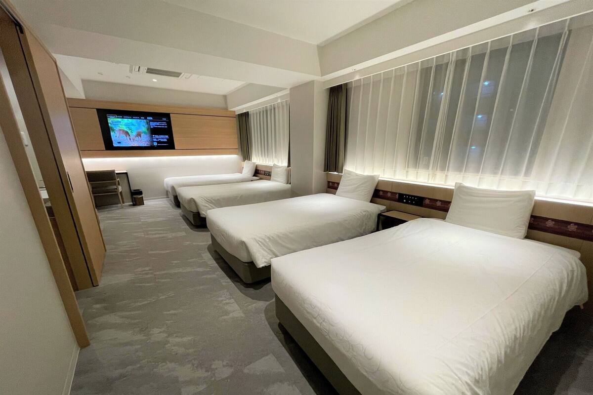 A hotel room with three beds, a TV mounted on the wall, and a large window with curtains.
