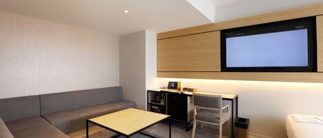A modern hotel room with a sofa, a small table, a desk with a computer, and a TV mounted on the wall.