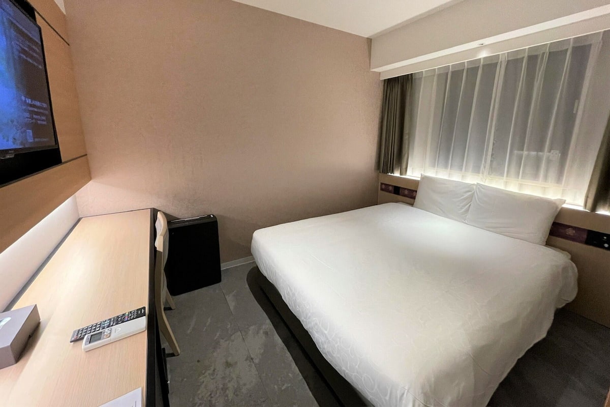 A hotel room with a bed, a TV mounted on the wall, and a desk with a remote control.
