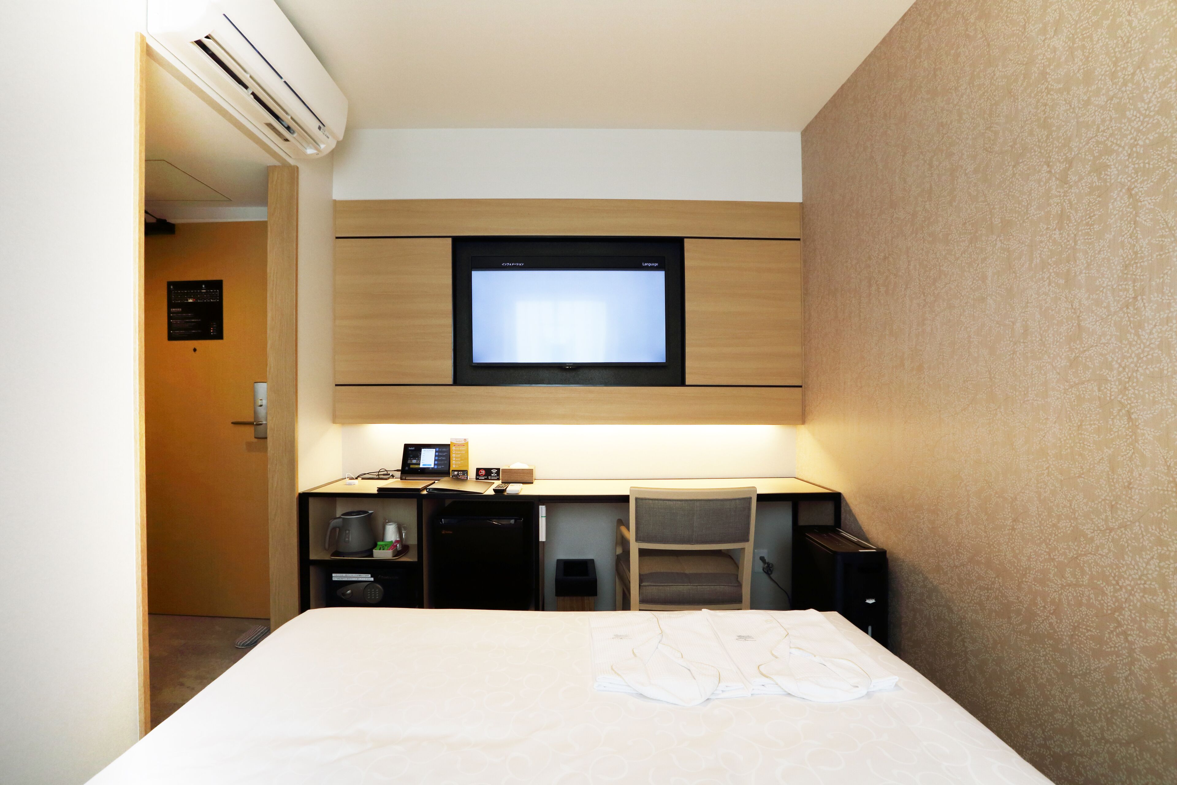 A hotel room with a bed, a desk with a computer, a chair, a TV mounted on the wall, and an air conditioning unit.