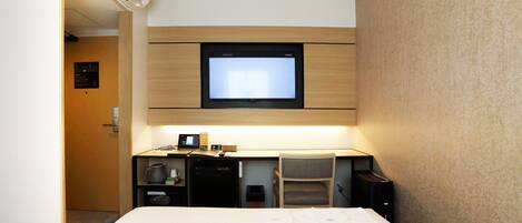 A hotel room with a bed, a desk with a computer, a chair, a TV mounted on the wall, and an air conditioning unit.