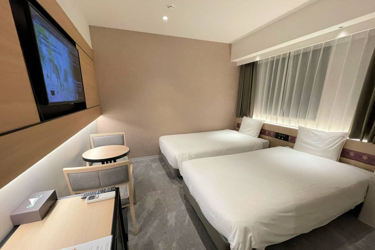 A hotel room with two beds, a small table with a chair, a TV mounted on the wall, and a window with curtains.