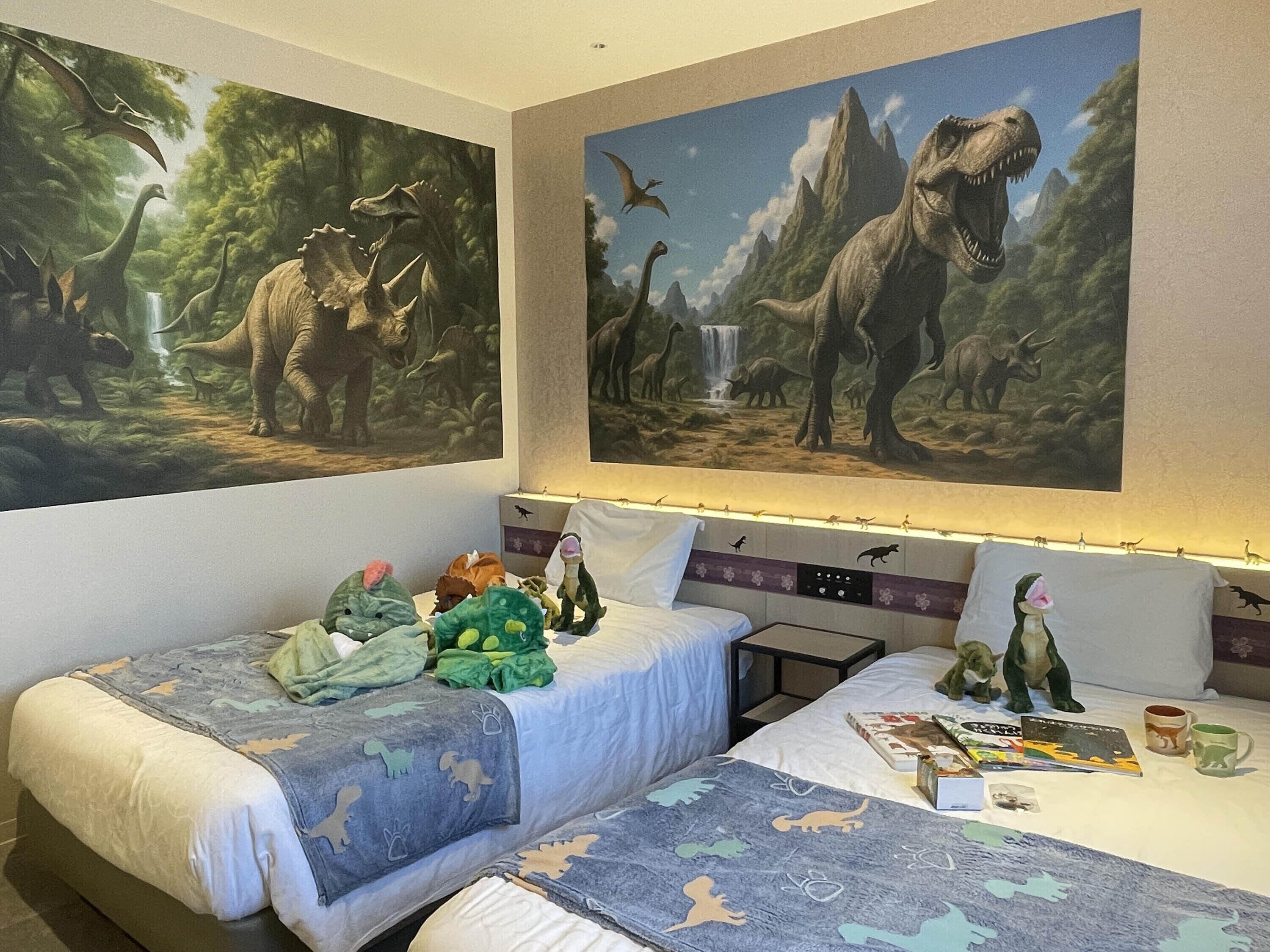 Quarto de hotel com duas camas, roupa de cama com tema de dinossauros e grandes murais de dinossauros nas paredes.